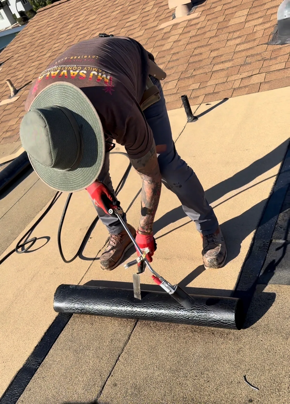 A worker wearing a wide-brimmed hat and red gloves uses a torch to apply a rolled black membrane onto a flat roof surface.