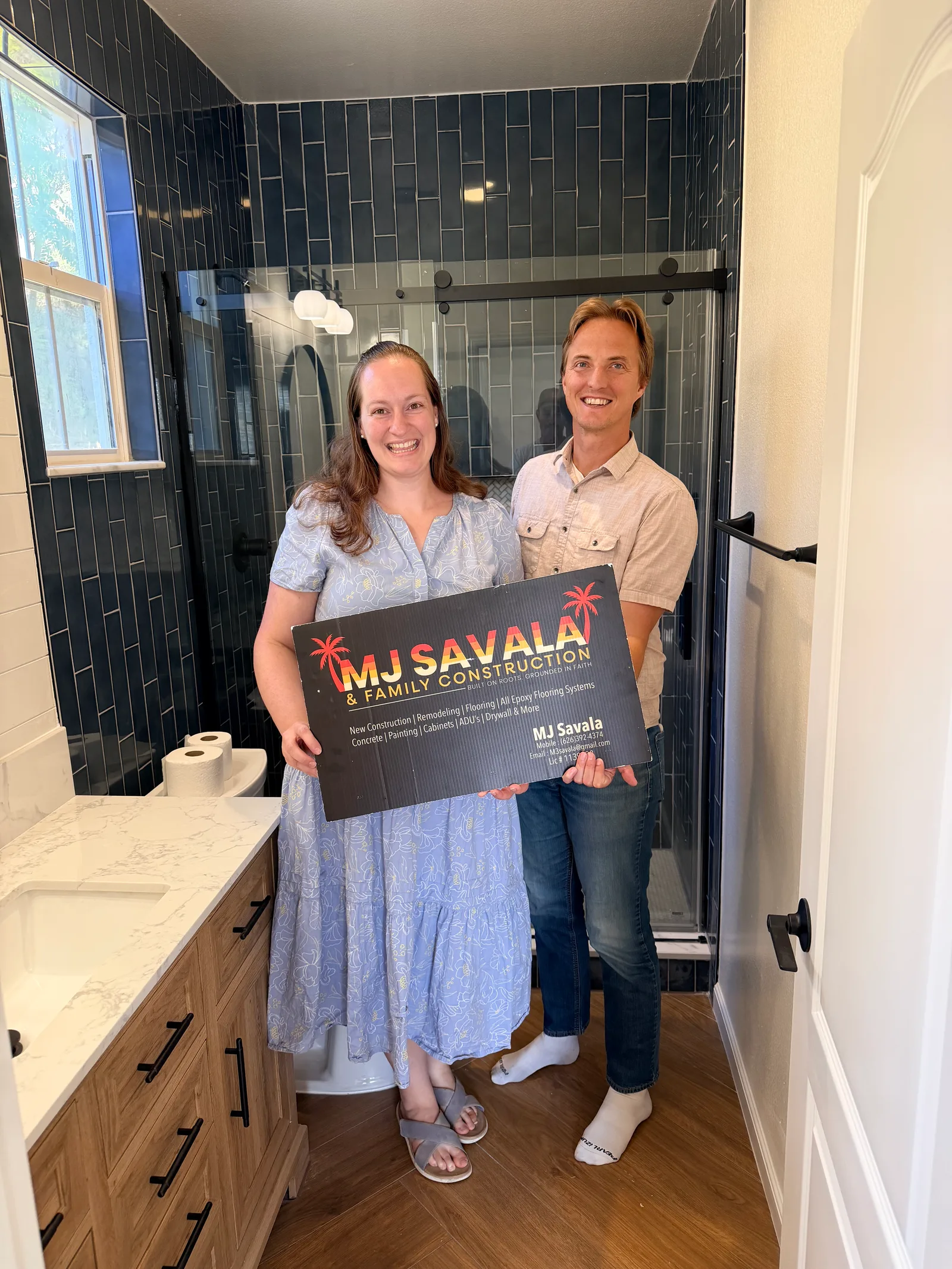 A man and woman stand smiling in a remodeled bathroom with dark tiled walls, a glass shower enclosure, a wood vanity with marble countertop, and wood-look flooring, holding a company sign.