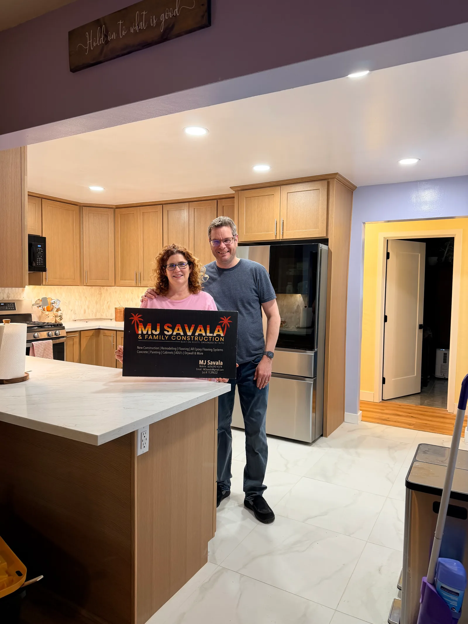 Two people stand in a remodeled kitchen with wood cabinets, a marble-topped island, tile flooring, and a stainless steel refrigerator, holding a business sign.